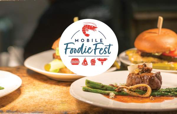 Mobile Foodie Fest logo (white circle with blue text and red icons of a shrimp, a burger, a pizza slice, a cupcake and a sausage on the end of a fork) with burgers and steak bites in the background