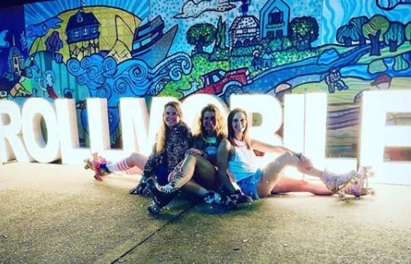 three women sitting on a sidewalk in front of 3 foot lighted letters reading # roll mobile