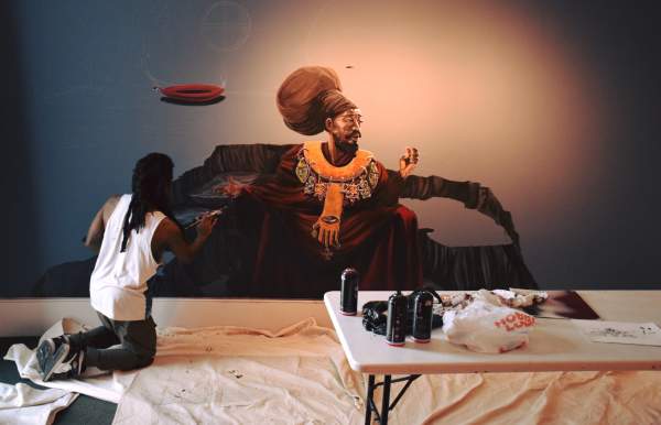 An artist kneels as he paints an African woman on a wall with artist supplies on a table