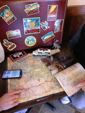 Two travelers plan a Wyoming road trip using a state map and Wyoming travel guide. Wyoming-themed stickers, a miniature truck and travel materials surround the map as one traveler points to a route, highlighting trip planning and exploration across the state.