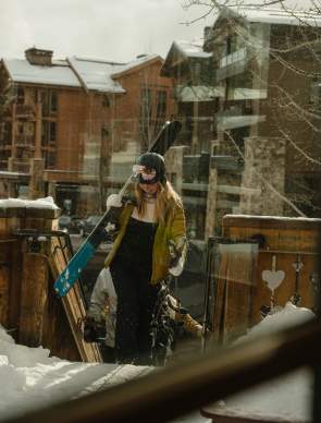 A skier carrying skis walks through deep snow toward a lodge after a day on the slopes near Jackson, Wyoming. Snow-covered buildings and a rustic wooden deck frame the winter scene, highlighting the après-ski experience and mountain lifestyle in Teton County.