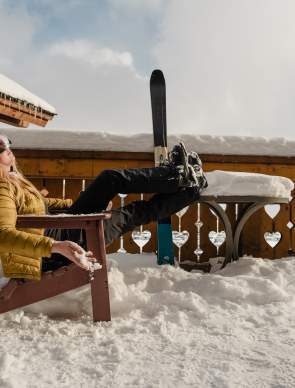 A skier relaxes in an outdoor chair surrounded by deep snow during an après-ski break near Jackson, Wyoming. Wearing ski goggles and winter gear, the skier rests their boots beside upright skis on a snow-covered deck, highlighting the relaxed side of a Wyoming winter adventure.