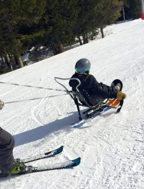 An adaptive skier uses a sit-ski while being assisted by an instructor on a snow-covered ski slope in Wyoming. The skier wears a helmet and winter gear as they navigate the mountain, highlighting accessible outdoor recreation and adaptive skiing opportunities.