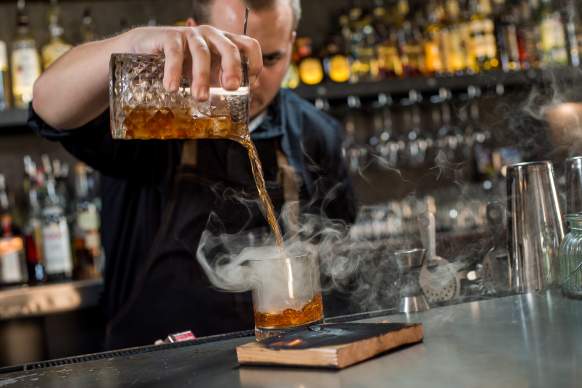 Bartender pouring a smoking Cedar cocktail at a bar with aromatic presentation