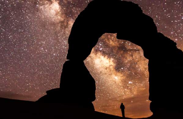 A solitary figure silhouetted beneath the iconic Delicate Arch in Arches National Park, Utah, framing the core of the Milky Way galaxy.