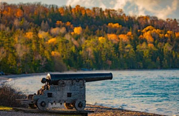 A cannon points over the water from the beach at Mackinac Island’s British Landing with fall color in the background