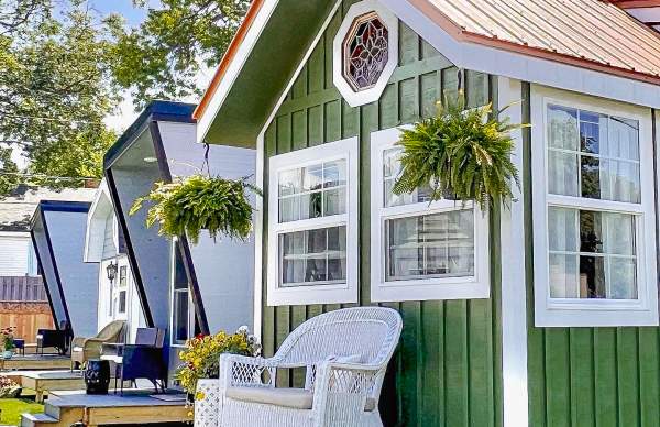 Green vertical-sided tiny house with a small front porch furnished with white wicker seating, set against a blue sky and leafy green tree.
