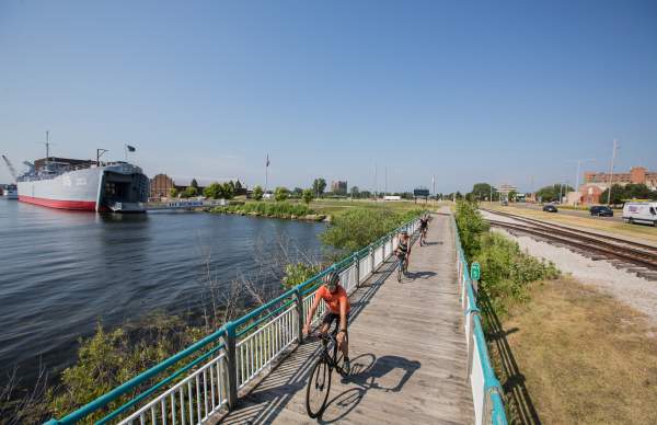 aeriel view of bikers on bike trail bridge going over water with WWII landing ship tank in background