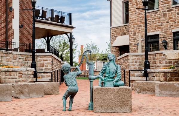 Bronze sculpture of a child pumping water for a seated Native American woman in a brick plaza surrounded by historic buildings.
