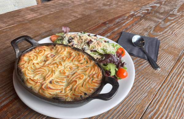 A plate featuring a shepherd's pie in a cast-iron dish with swirled mashed potato topping, served alongside a mixed green salad with cherry tomatoes, almond slivers, and shredded cheese on a rustic wooden table.