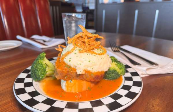 A plated meatloaf dish from Cap City, featuring a slice of meatloaf topped with mashed potatoes, crispy fried onions, and garnished with parsley. The dish is served with broccoli florets and surrounded by a vibrant orange sauce on a black-and-white checkered plate, set on a wooden table in a diner-style setting.