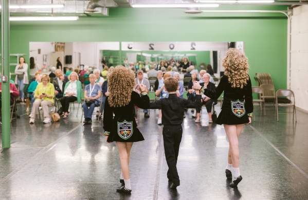 Three young Irish dancers perform for a seated audience, wearing black costumes with colorful embroidered patches.