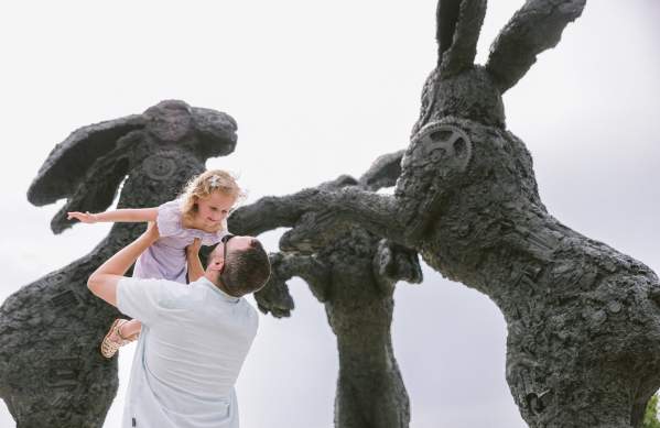 A father lifting his smiling daughter in front of large, textured rabbit sculptures in a whimsical outdoor setting, under a bright sky.