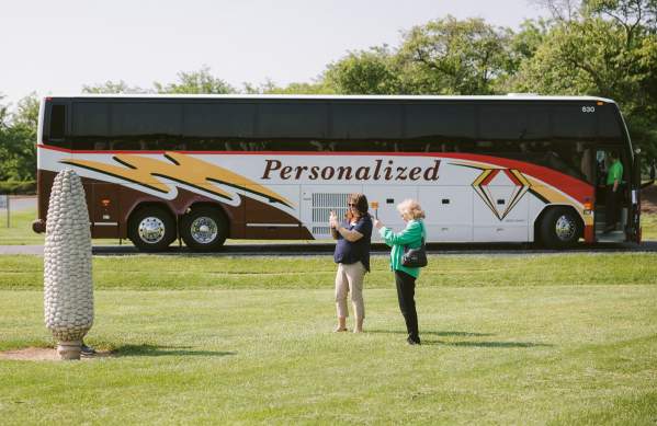 A group tour motorcoach parked at the Field of Corn art installation with visitors taking photos in the field