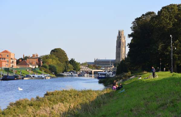 A cycle path srurronded by grass and trees next to a river. Boston Stump and town can be seen in the distance.