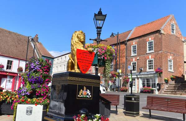 Golden lion water pump on a black plinth with a red commemorative shield, surrounded by colourful flower displays, set in a Caistor Market Place, with red-brick buildings and a lamppost under a clear blue sky.