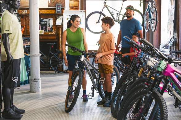 A family interacts in a bike shop, surrounded by various mountain bikes and cycling gear. The shop features a rustic interior with bikes displayed on walls.
