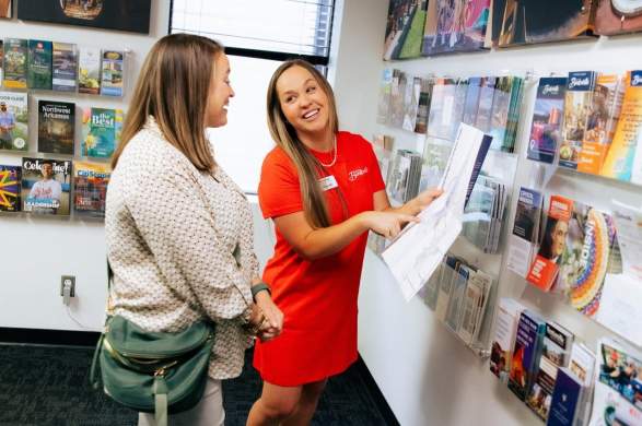 A woman in a red dress assists another woman in a casual outfit, discussing brochures in a well-lit visitor center. Informative displays line the walls.