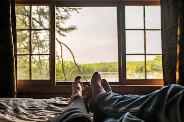 Two people's feet with the window showing view on the lake/wilderness from their cabin