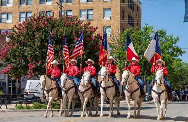 Image of 6 Women in hot pink shirts riding through downtown on 6 white horses with three holding American Flags and the other three holding three texas flags.