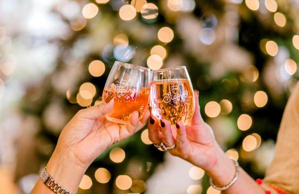 Two women tapping two wine glasses together for a cheers to the holidays in front of a lighted christmas tree in the background.