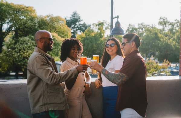 Four adults perched on a rooftop patio cheers their beverages with smiles and a happy atmosphere.