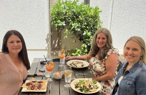three ladies sit at a table outdoors with drinks and plates with food