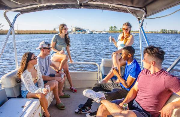 Six young people seated on a covered pontoon boat with Tchefuncte River and shoreline behind.