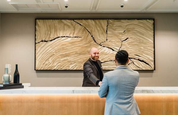 Man in a black suit behind a hotel check-in desk smiles and reaches towards the man on the other side of the desk who is seen from the back.
