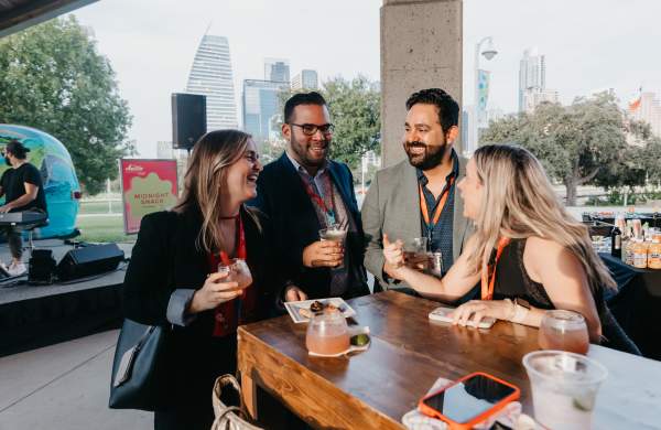 4 people wearing conference badges stand together at a tall wooden table during an outdoor event on the Palmer Event Center with the Austin Skyline in the background and cocktails in hand