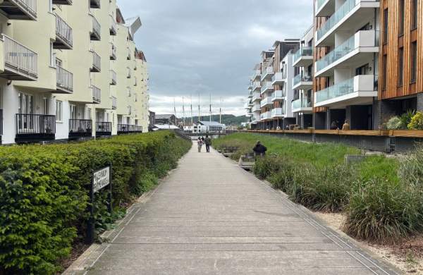 An image of a wide pathway with neat green bushes running along either side. There are tall buildings on either side that are light yellow and clad in wood. In the distance there are two people walking ahead and even further there are six masts of a ship.