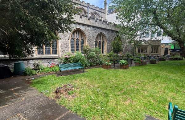 An image of a grassy lawn area surrounded by narrow paved paths. There is a church behind the path and benches and flowers pots on the path running alongside the church.