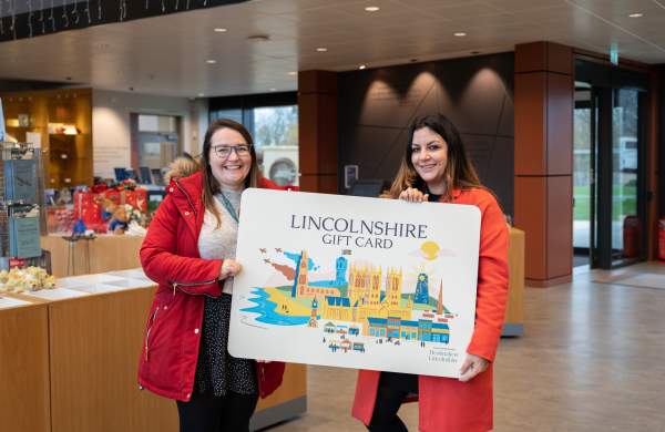two women in red coats holding an oversized 'Lincolnshire Gift Card' inside the visitors centre at IBCC