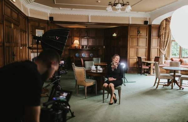 A woman sat on a chair in the middle of a hotel lounge with camera and a cameraperson in the foreground