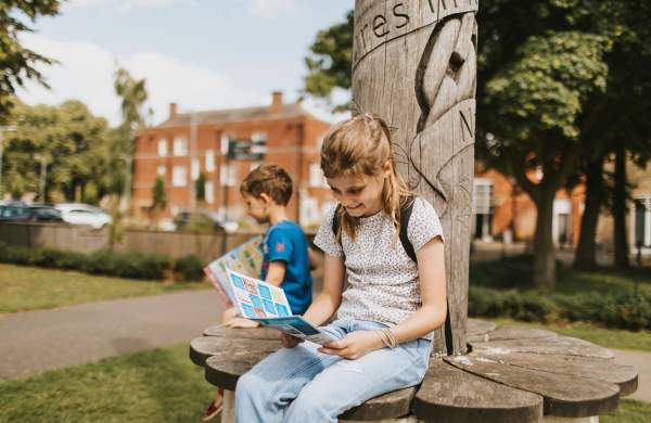 Girl looking at a treasure trail map sat on a bench with young boy in background