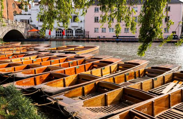 A row of 12 punts on the river in Cambridge with a stone bridge to the left & a white & pink building on the other side of the river