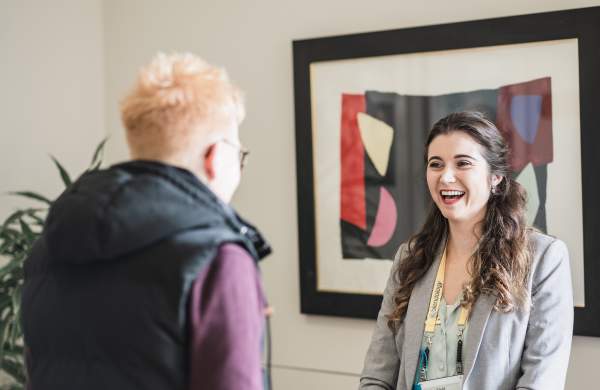 A young white woman with long wavy brown hair smiling in front of an abstract art picture whilst chatting to another white woman who has her back to the camera
