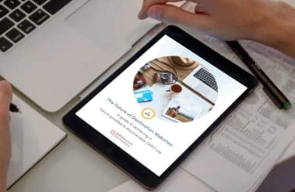 A tablet sitting on a desk by a laptop and some papers, open to the first page of a Granicus Destinations guide titled, "The future of destination website starts here."