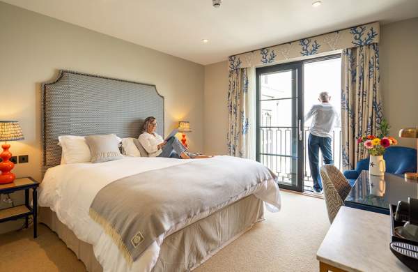 A man and a woman in a hotel room. Woman reading on the bed, man looking out of the window.