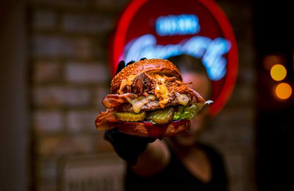 A close up of a man holding a burger up to camera in a dim lit restaurant