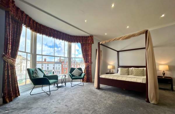 a bed and two chairs in a hotel room by a bay window overlooking a town square