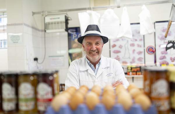 A man in butchers clothing looking at camera surrounded by butchers products