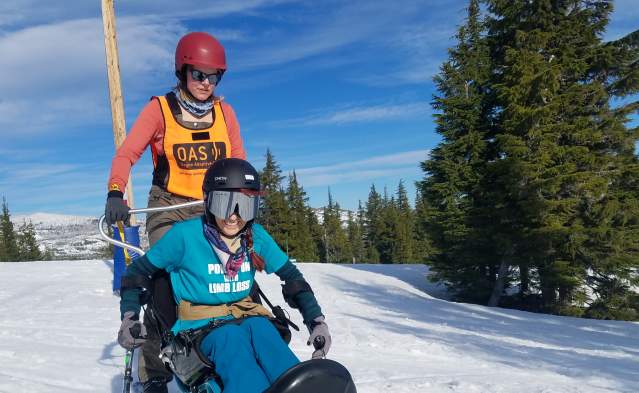 A skier uses adaptive ski equipment with a companion to ski the slopes. Snow, trees and blue sky in the backdrop.