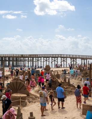 People walking around multiple sand sculptures on the beach during the sandcastle days festival.