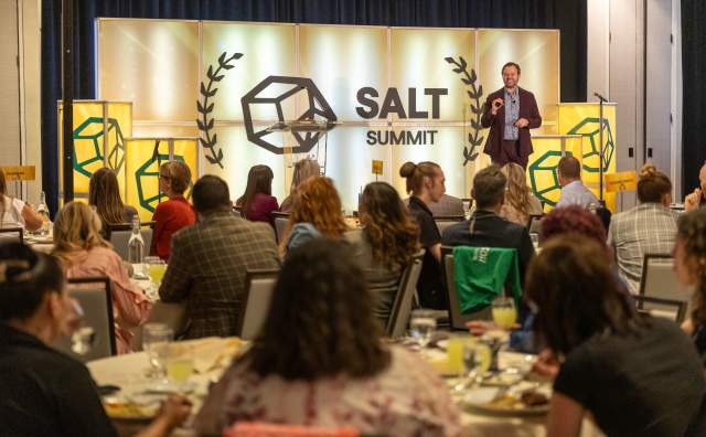 Crowd at tables listening to a speaker on stage. The stage says Salt Summit with laurel wreaths around it