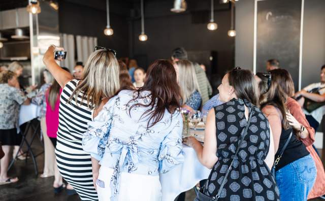 5 women standing together around a tall table, backs to you they are taking a group photo selfie