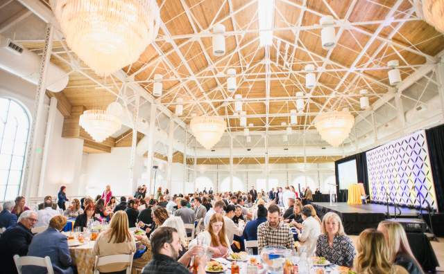 Warmly lit large room filled with people sitting at round tables and a speakers stage at the front.