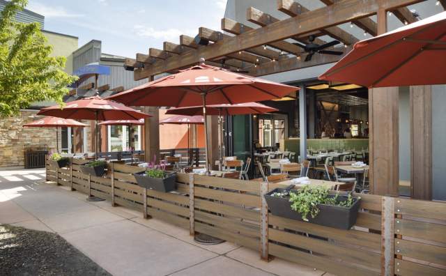 Outdoor patio that has tables with reddish sun-shade umbrellas looking into the restaurant at Hearth and Hill