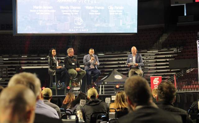 Panel of 2 men and one woman on stage with a moderator asking questions and a presentation on the screen above and behind them