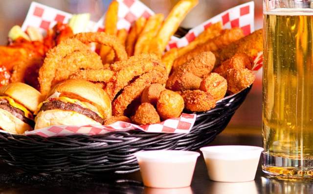 Basket of onion rings, fries, mozzerella sticks, sliders with a stein of beer beside it and containers of dipping sauces in front